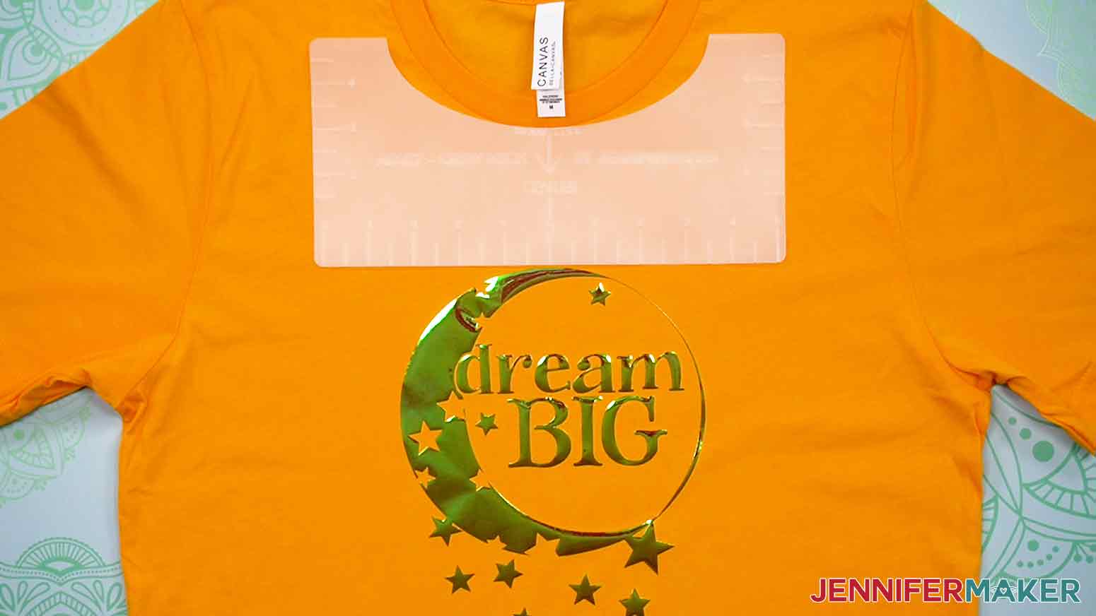 Another example of an orange, adult crew-neck T-shirt with the plastic Adult Crew-Neck T-Shirt Ruler aligned under the neckline of the shirt shows the proper placement and spacing of the Dream Big decal.