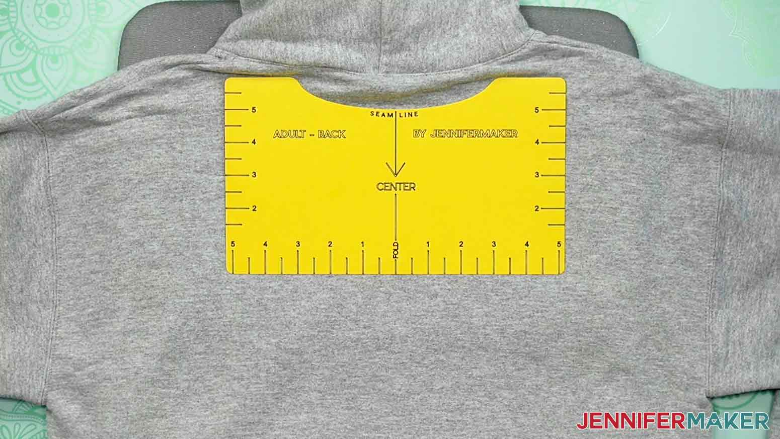 Here is the cardstock Adult Back T-Shirt Ruler placed under the neckline seam on the back of a grey adult hoodie, showing that it is the correct size to be used as a guide for the back of an adult hoodie.