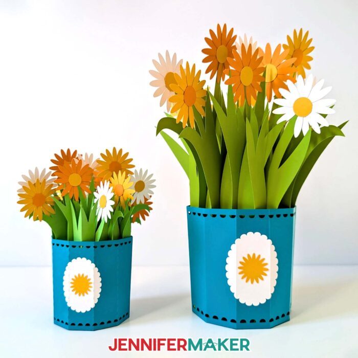 Two matching 3D Pop-Up Spring Flowers bouquets with blue pots and a mixture of yellow and white daisies. One pot is a small version cut with a Cricut Joy, and the other is the full-size version for other cutting machines