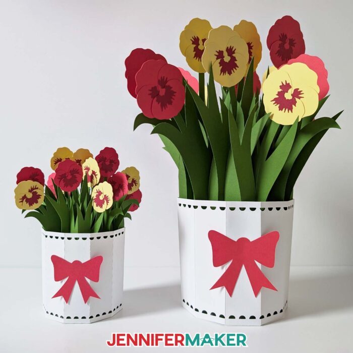 Two matching bouquets of 3D Pop-Up Spring Flowers with white pots and red bows, with red and yellow pansies inside. One pot is a small version cut with a Cricut Joy, and the other is the full-size version for other cutting machines