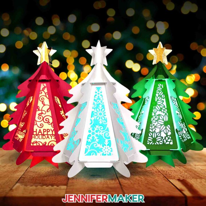 Learn how to make a 3D paper tree lantern with JenniferMaker's tutorial! Three cardstock trees sit illuminated against a background lit with Christmas lights.