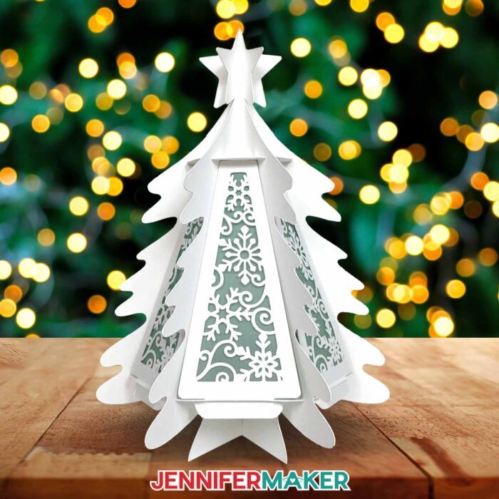Learn how to make a 3D paper tree lantern with JenniferMaker's tutorial! A cardstock tree sit illuminated against a background lit with Christmas lights.