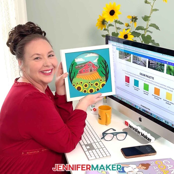 Jennifer Maker sits at her desk in a red blouse in front of her computer, smiling as she shows off a barn shadow box using the color palette shown on her computer screen in Canva from her Best Canva Tools For Business post