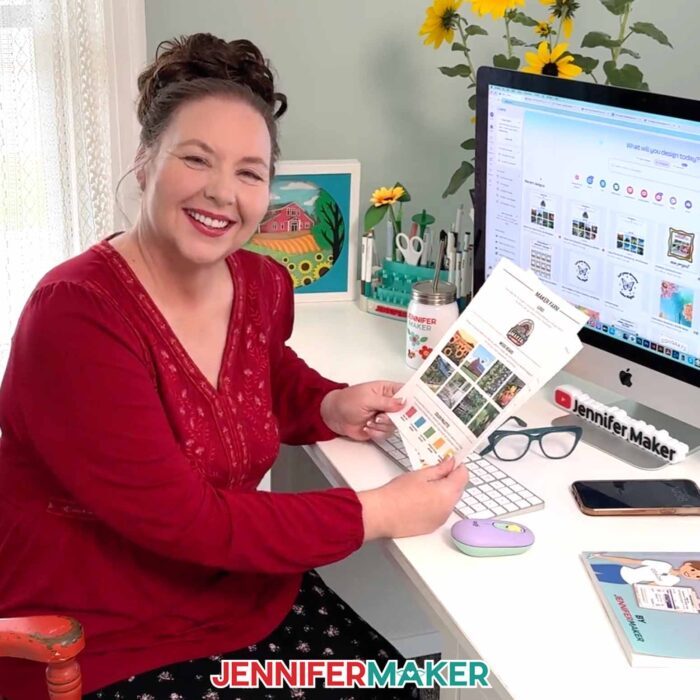 Jennifer Maker sits at her desk in a red blouse in front of her computer, smiling as she shows off some printed pages from her Maker Marketplace book, in front of her computer screen pulled up to Canva, ready to share her Best Canva Tools For Business