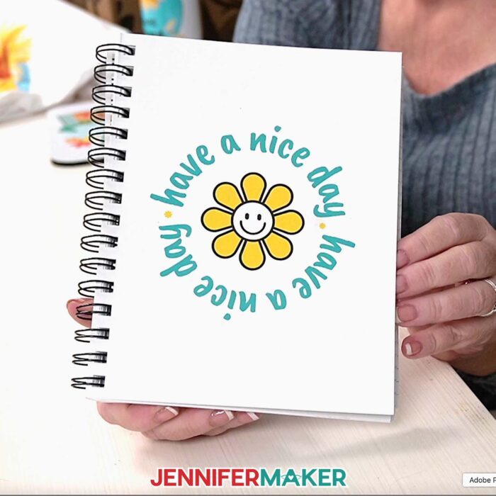 Jennifer holds up a notebook with a smiling cartoon sunflower and the words "Have a nice day" printed on the cover.