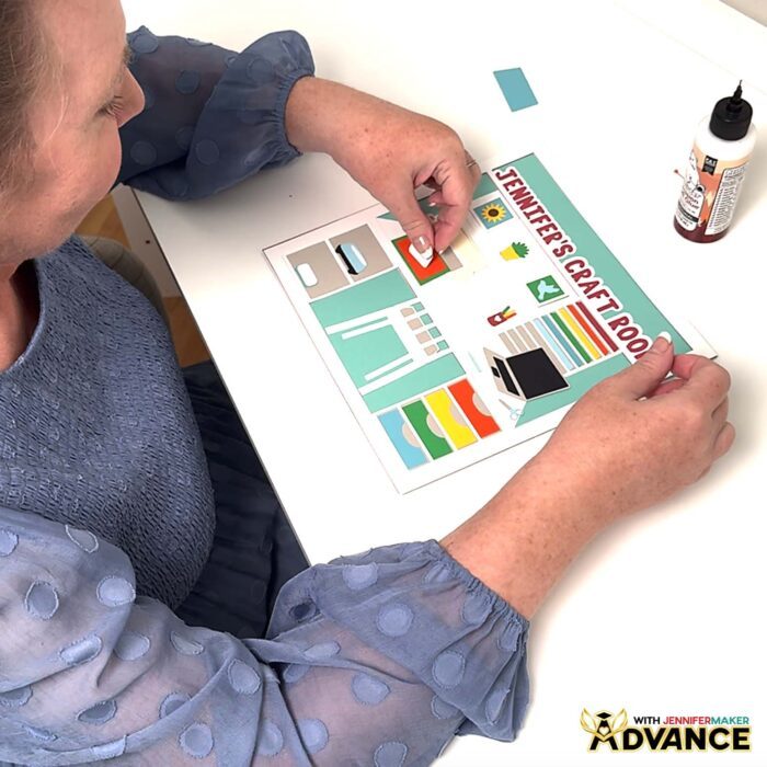 Jennifer Maker sits at her desk and assembles a custom multi-layered craft room shadow box design with "Jennifer's Craft Room" added to the top