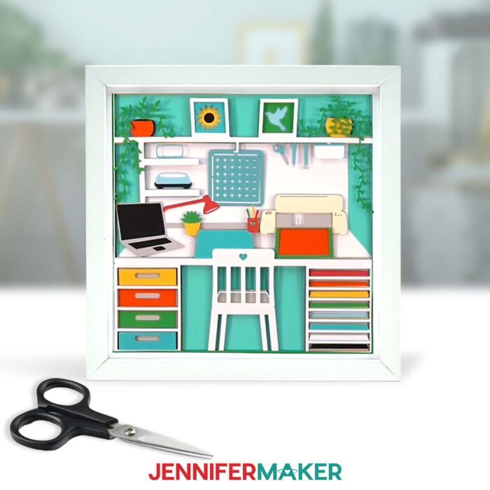 A colorful craft room shadow box design sits on display in a white frame, with a pair of black handled scissors in front