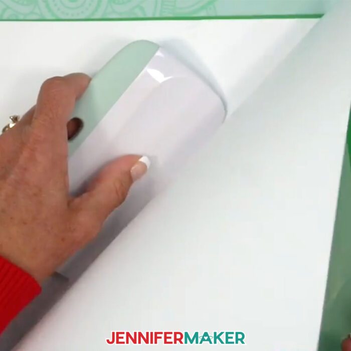 Jennifer Maker demonstrates how to use the Cricut XL scraper tool to apply white Cricut Value Vinyl to a piece of cardstock to reinforce it for cleaner intricate cuts in the craft room shadow box design tutorial