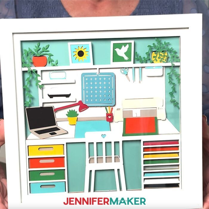 Jennifer Makers holds and shows off the details of her craft room shadow box design, with colorful tools, drawers, and a cute white chair at the desk