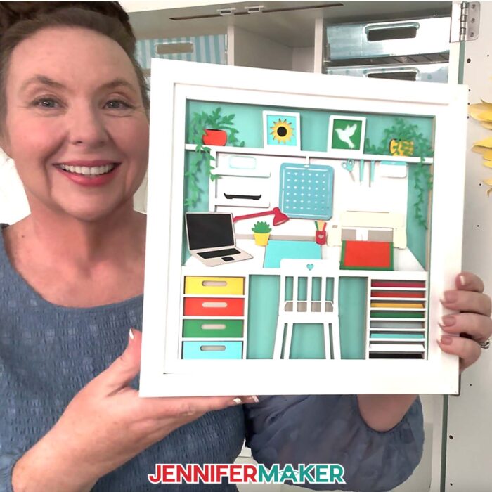 Jennifer Maker holds and shows off her craft room shadow box design, full of colorful details, in a white frame