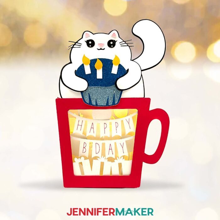 A red Hot Chocolate Shadow Box, with a smiling cardstock cat holding a glittery blue cupcake with three candles. Inside the shadow box is a banner reading "Happy B-Day"