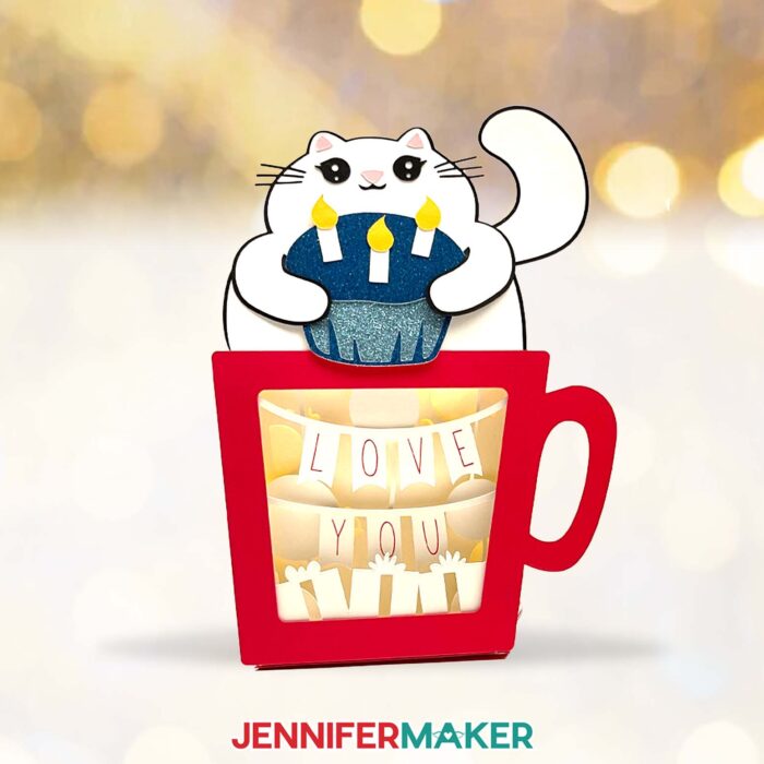 A red Hot Chocolate Shadow Box, with a smiling cardstock cat holding a glittery blue cupcake with three candles. Inside the shadow box is a banner reading "Love You"