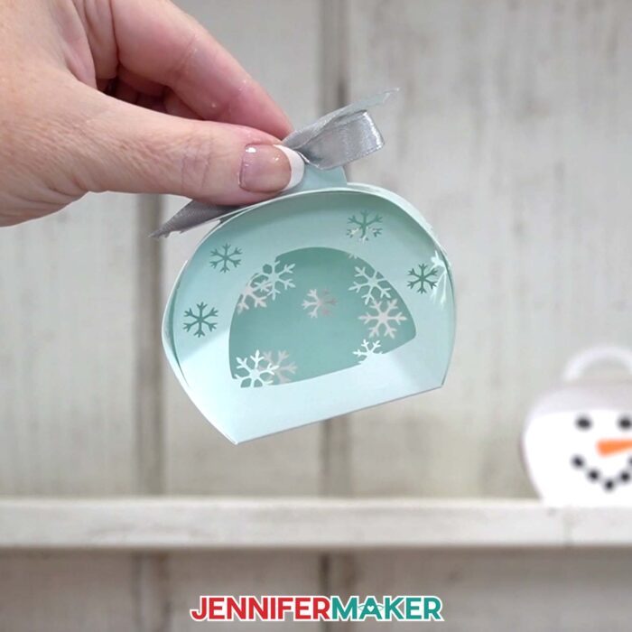 A hand shows off the snowflake box from the Curvy Keepsake Boxes tutorial, with the snowman box in the background