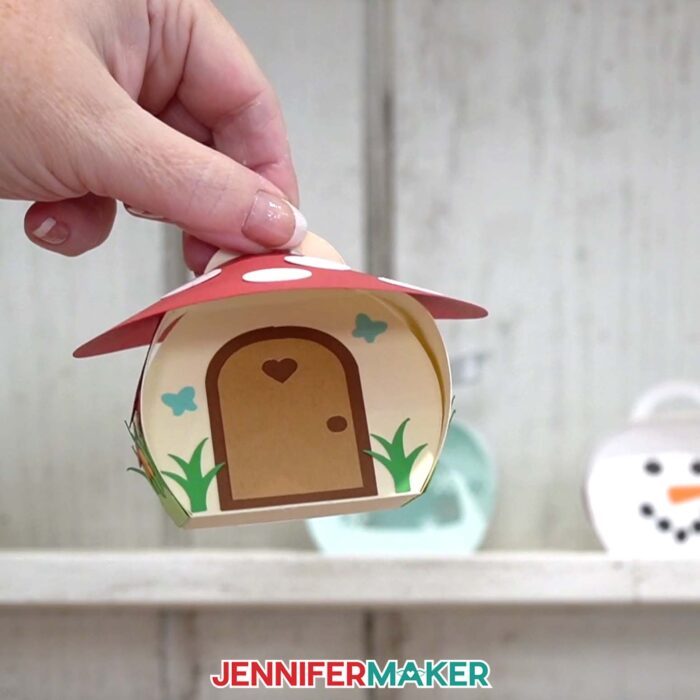 A hand shows off the mushroom house box from the Curvy Keepsake Boxes tutorial, with the snowflake and snowman boxes in the background
