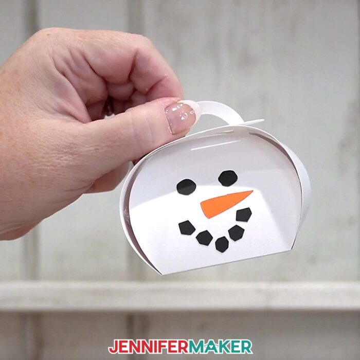 A hand shows off the snowman box from the Curvy Keepsake Boxes tutorial, against a whitewashed fence