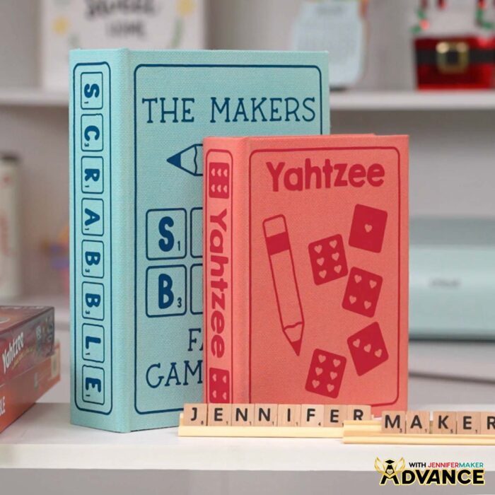 Learn how to customize your DIY Board Game Box through the Advance with JenniferMaker program! A large, blue painted book box labeled "Scrabble" on the spine and "The Makers" on the cover stands beside a medium, red painted box labeled "Yahtzee" on the front and spine. A rack of letter tiles in front spells out "Jennifer Maker."