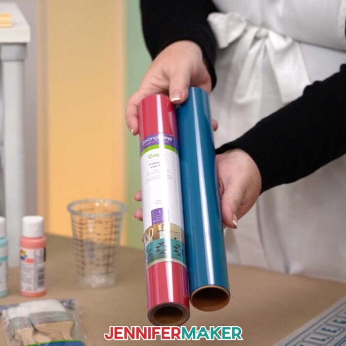 Learn how to make a DIY Board Game Box to store all your game pieces with JenniferMaker's tutorial! Jennifer holds up a roll of red heat transfer vinyl and a roll of blue heat transfer vinyl.