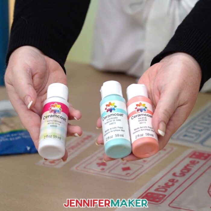 Learn how to make a DIY Board Game Box to store all your game pieces with JenniferMaker's tutorial! Jennifer holds a bottle of textile medium in one hand and bottles of blue and red acrylic paint in the other hand.