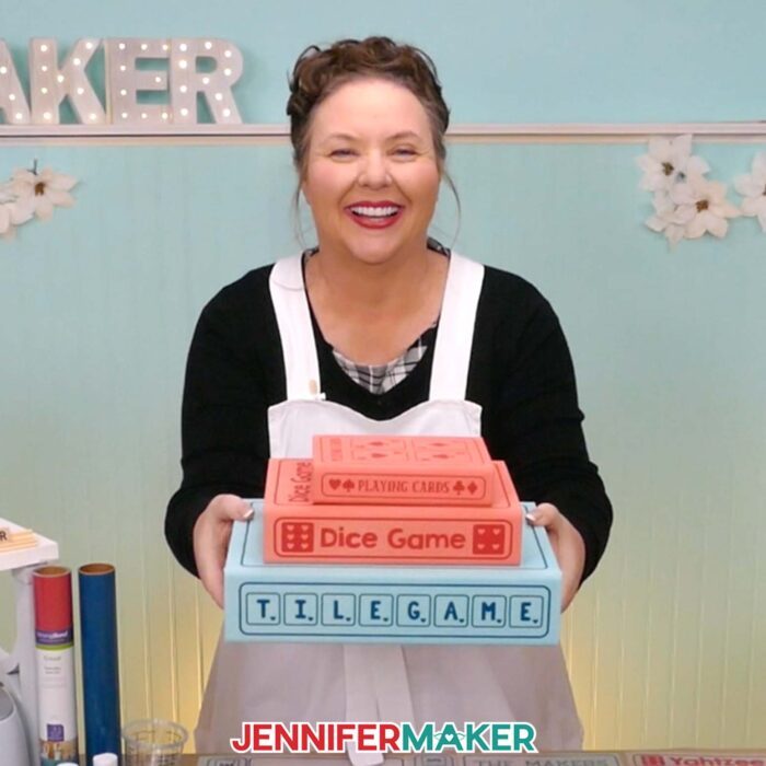 Learn how to make a DIY Board Game Box to store all your game pieces with JenniferMaker's tutorial! Jennifer holds a stack of three painted book boxes with iron-on vinyl labels. A small red box labeled "Playing Cards" is on top, a medium red box labeled "Dice Game" is in the middle, and a large blue box labeled "Tile Game" is on the bottom.