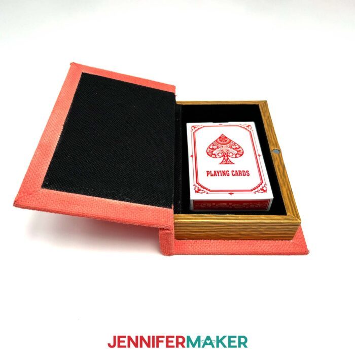 Learn how to make a DIY Board Game Box to store all your game pieces with JenniferMaker's tutorial! A small, painted red book box lies open with a deck of playing cards inside.