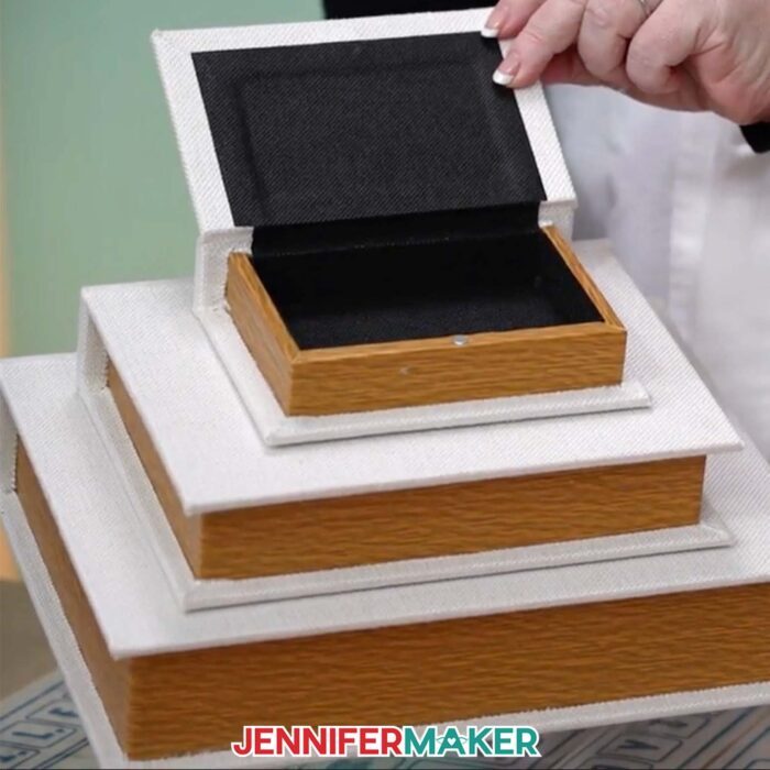 Learn how to make a DIY Board Game Box to store all your game pieces with JenniferMaker's tutorial! Three white book boxes are arranged in a stack. A large box is on the bottom, a medium box is in the middle, and a small, open box is on top.