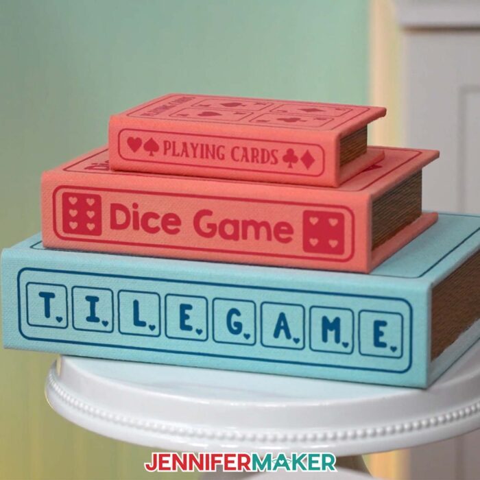Learn how to make a DIY Board Game Box to store all your game pieces with JenniferMaker's tutorial! Three painted book boxes with iron-on vinyl labels are stacked on a stool. A small red box labeled "Playing Cards" is on top, a medium red box labeled "Dice Game" is in the middle, and a large blue box labeled "Tile Game" is on the bottom.