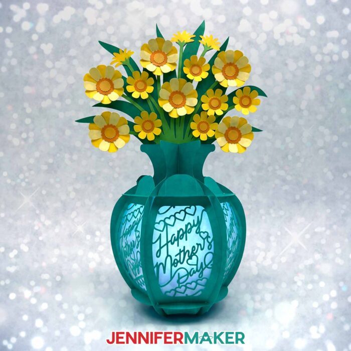 A teal cardstock lantern vase with panels that say Happy Mother's Day". A bouquet of yellow and white flowers rest in the vase. Flower Bouquet Lantern design by JenniferMaker.