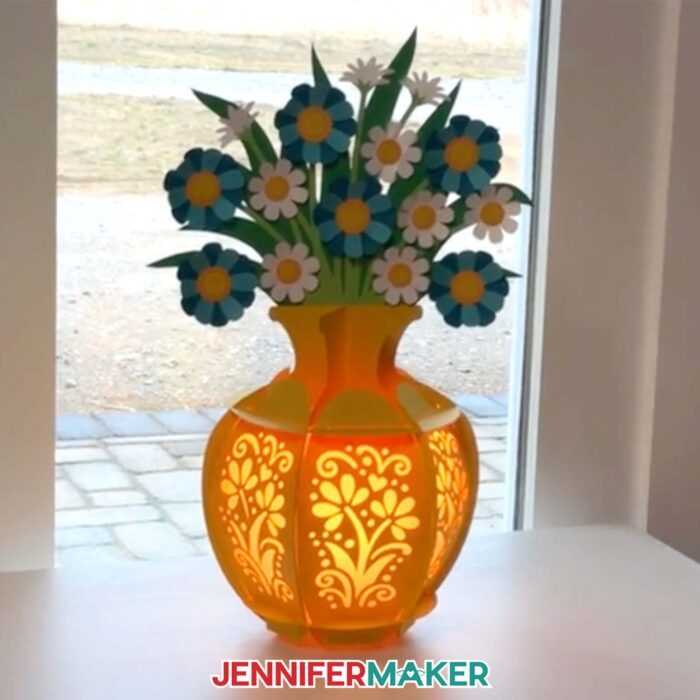 A yellow lantern vase with a blue and white flower bouquet is lit up on a table. Flower bouquet lantern by JenniferMaker.