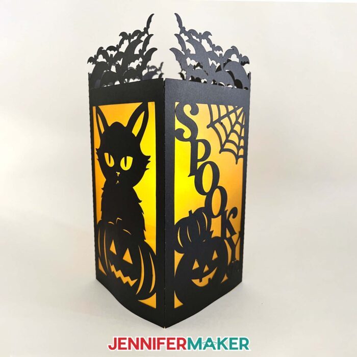 Black Halloween paper lantern showing the two panels with a cat, pumpkins, "spooky,"  and bats with orange vellum diffusing the light inside.