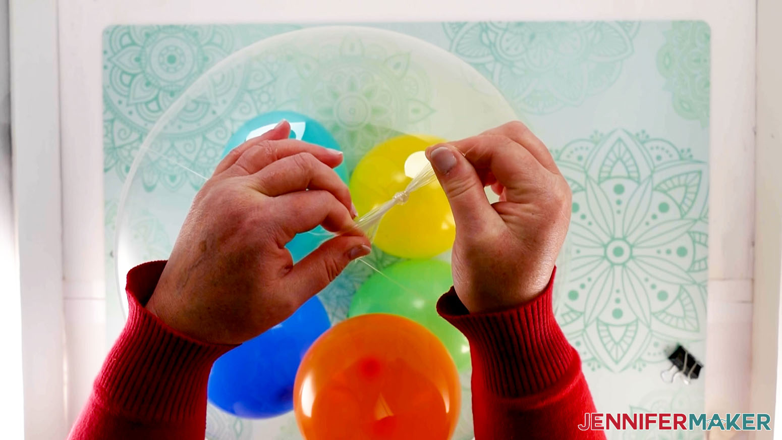 An overhead photo showing two hands tying a knot to secure an inflated Bobo balloon containing five smaller colored balloons