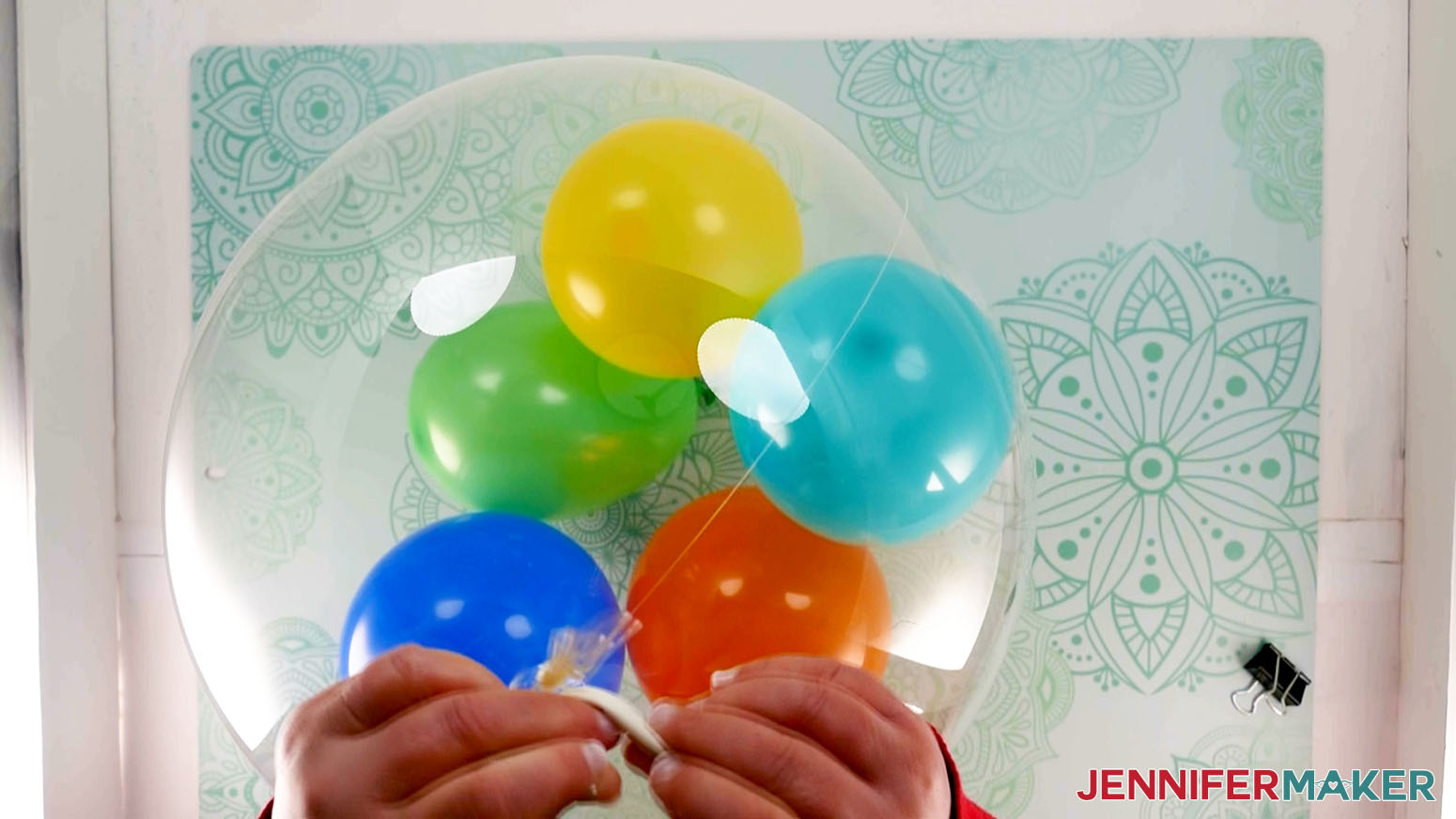 An overhead photo showing an uninflated white latex balloon being tied around the knot of an inflated Bobo balloon to secure the knot