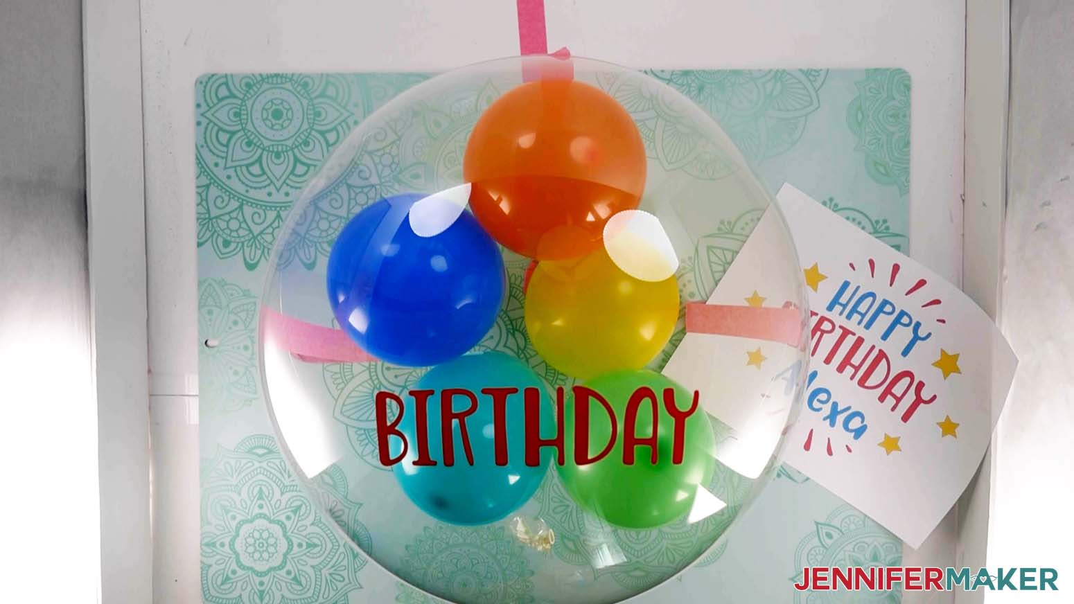 An overhead photo showing the word Birthday in red vinyl applied to an inflated Bobo balloon. A printout showing the entire vinyl balloon bouquet decal is lying on the work surface below.