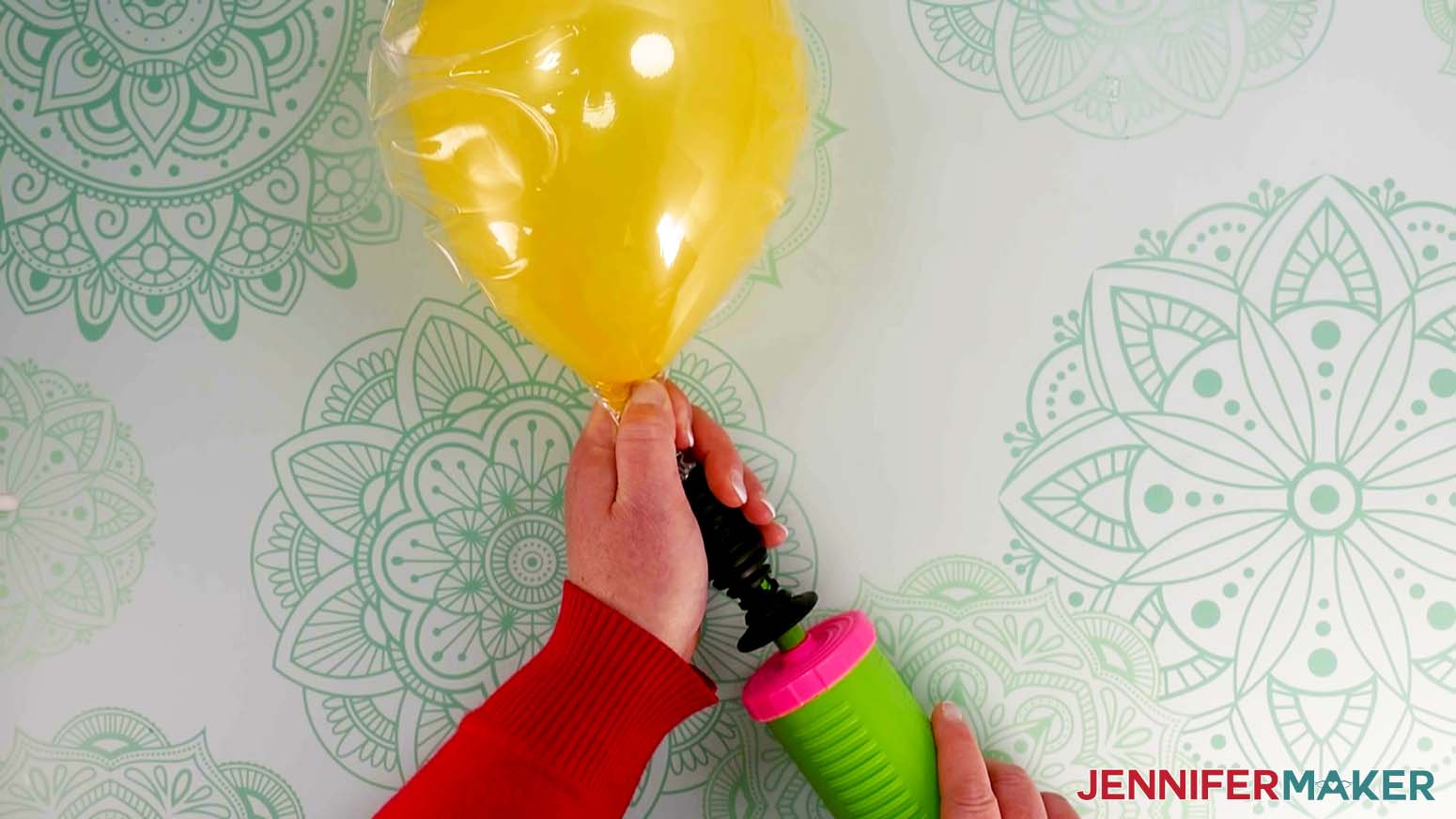 An overhead photo showing a yellow latex balloon attached to a hand pump, which is being used to inflate the balloon while inside of a deflated Bobo balloon