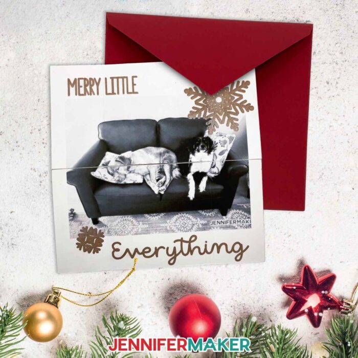 Make a custom infinity card with JenniferMaker's tutorial! An infinity card with a photo of her dogs Hunter and Chloe relaxing on a couch, with the sentiment "Merry Little Everything" written in copper pen with snowflakes sits on a speckled surface with a maroon envelope, some Christmas ornaments, and holiday greenery.