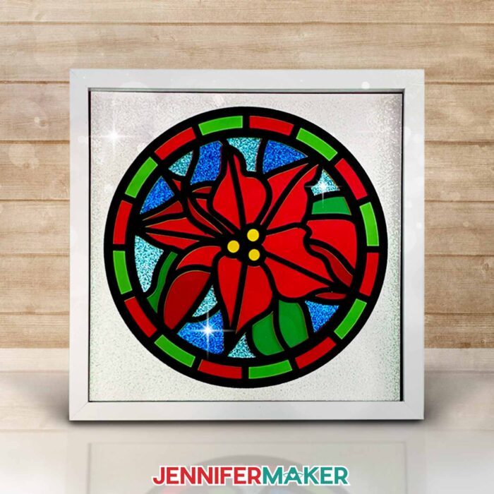 Make layered paper stained glass windows with Jennifer Maker's tutorial! Image shows a colorful poinsettia stained glass shadowbox in a white frame.