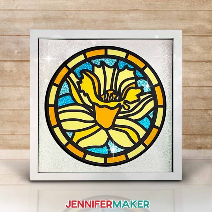 Make layered paper stained glass windows with Jennifer Maker's tutorial! Image shows a colorful daffodil stained glass shadowbox in a white frame.