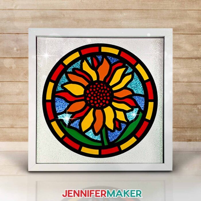 Make layered paper stained glass windows with Jennifer Maker's tutorial! Image shows a colorful sunflower stained glass shadowbox in a white frame.