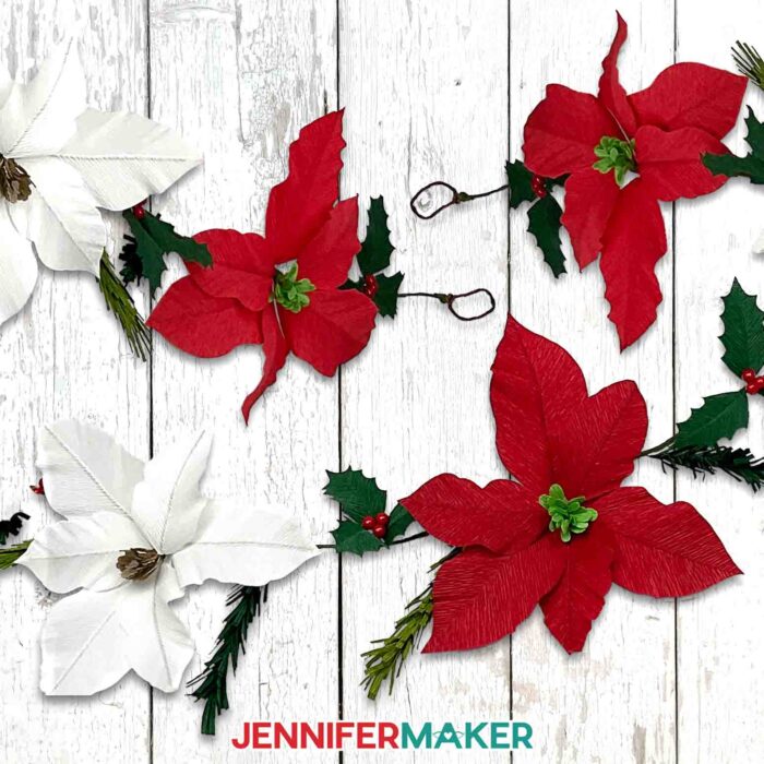 Learn to make a crepe paper floral garland with JenniferMaker's tutorial! A crepe paper garland of poinsettias, holly, and pine sprigs lays displayed across a painted white wooden surface.