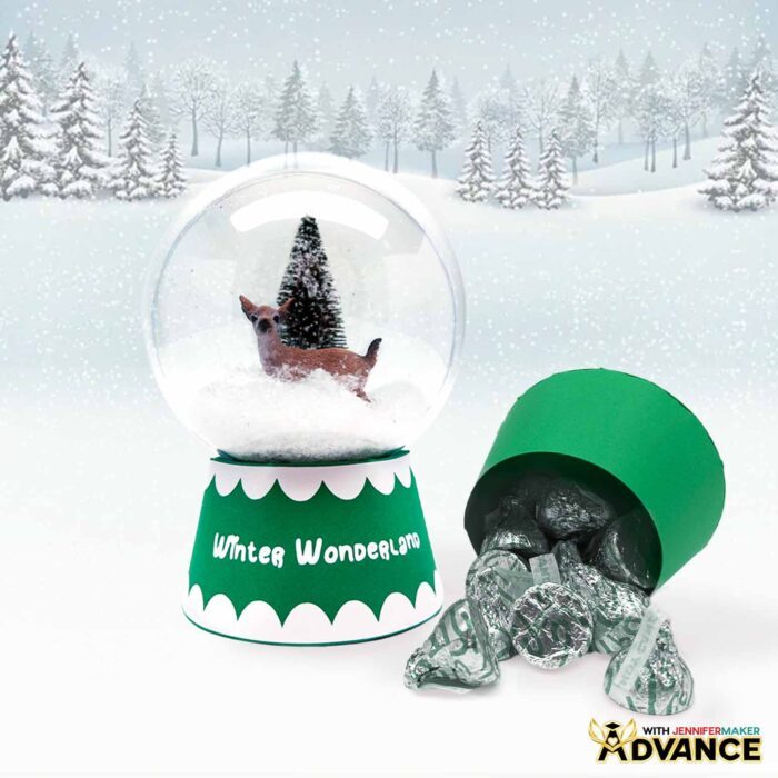 A green Snow Globe Gift Box design, with a tree and a deer inside sit on display on a snowy forest backdrop.