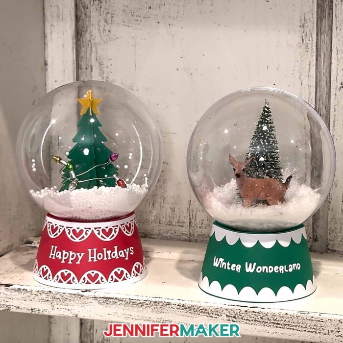 Two Snow Glob Gift Box projects sit next to each other on a shelf, one filled with a Christmas tree with lights, and one filled with a mini pine tree and a deer