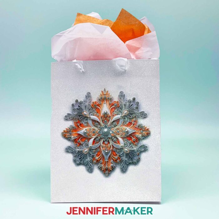 Make DIY Sublimation Gift Supplies with JenniferMaker's tutorial! A glitter gift bag stuffed with colorful tissue paper features an intricate sublimated snowflake design in orange tones.