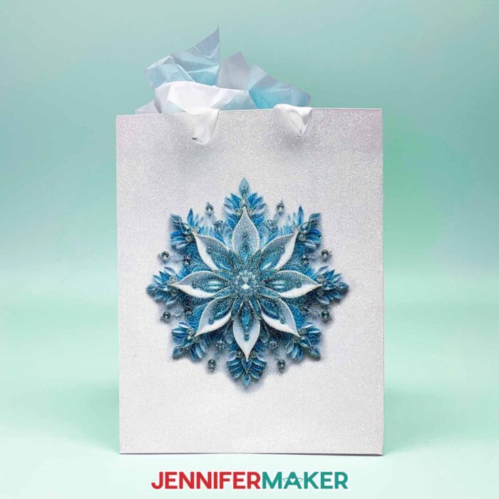 Make DIY Sublimation Gift Supplies with JenniferMaker's tutorial! A glitter gift bag stuffed with colorful tissue paper features an intricate sublimated snowflake design in blue tones.