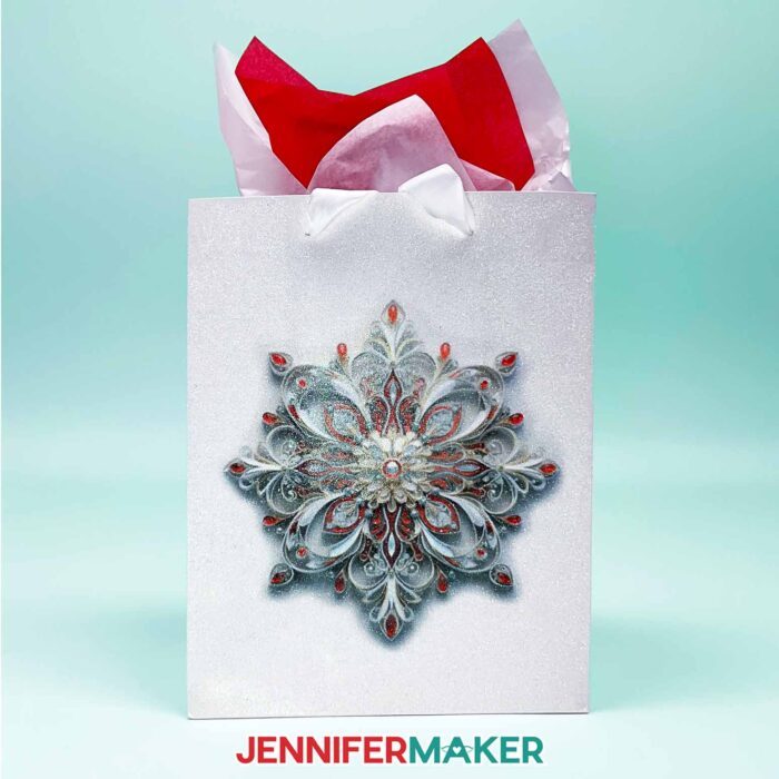 Make DIY Sublimation Gift Supplies with JenniferMaker's tutorial! A glitter gift bag stuffed with colorful tissue paper features an intricate sublimated snowflake design in red and gray tones.