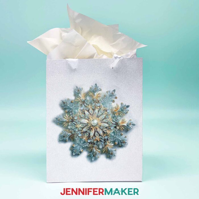 Make DIY Sublimation Gift Supplies with JenniferMaker's tutorial! A glitter gift bag stuffed with white tissue paper features an intricate sublimated snowflake design in soft blue tones.