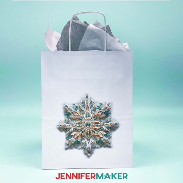 Make DIY Sublimation Gift Supplies with JenniferMaker's tutorial! A white gift bag stuffed with gray and white tissue paper features an intricate sublimated snowflake design in soft turquoise tones.