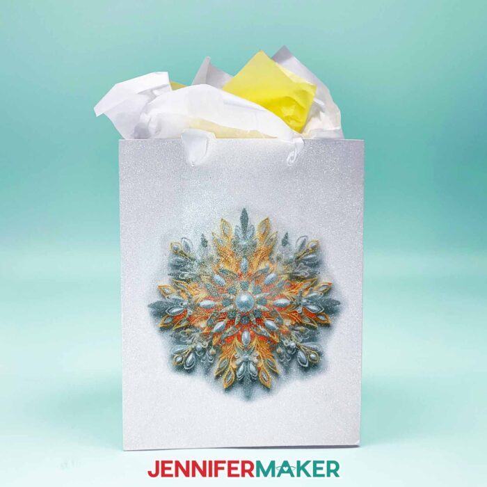 Make DIY Sublimation Gift Supplies with JenniferMaker's tutorial! A glitter gift bag stuffed with colorful tissue paper features an intricate sublimated snowflake design in yellow and gold tones.