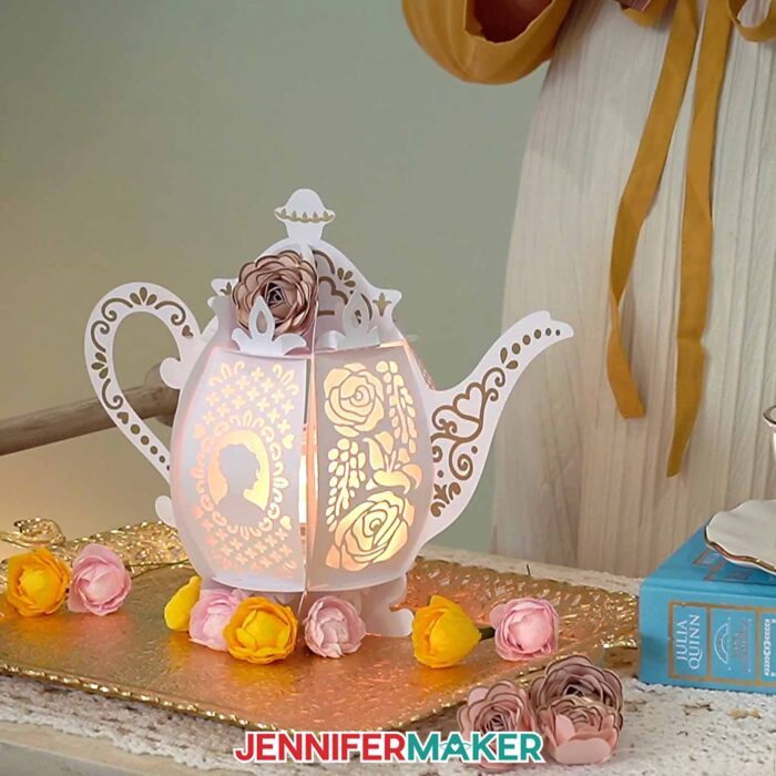 The lit-up teapot lantern rests on a tea tray with orange and pink flowers with a Bridgerton novel nearby.