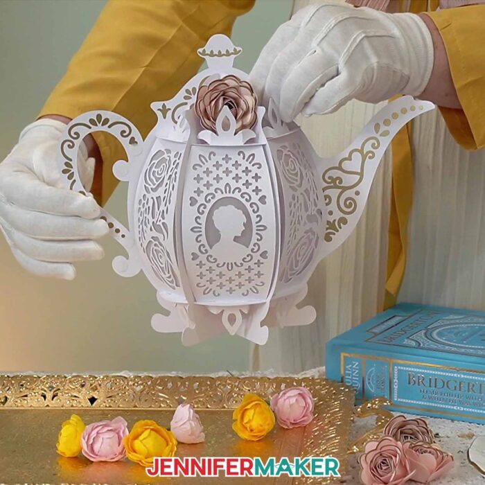 Jennifer's gloved hands hold up a tea-pot shaped lantern with intricate vinyl details and accent flowers.