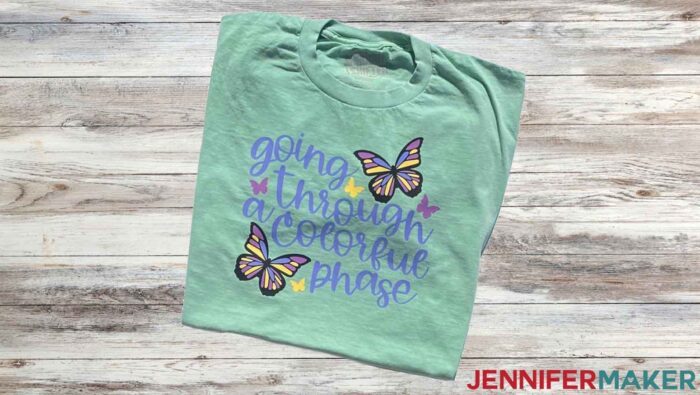 Green t-shirt with butterfly design turned blue, pink, and yellow in the sunlight. Learn to use color changing HTV with JenniferMaker's tutorial!