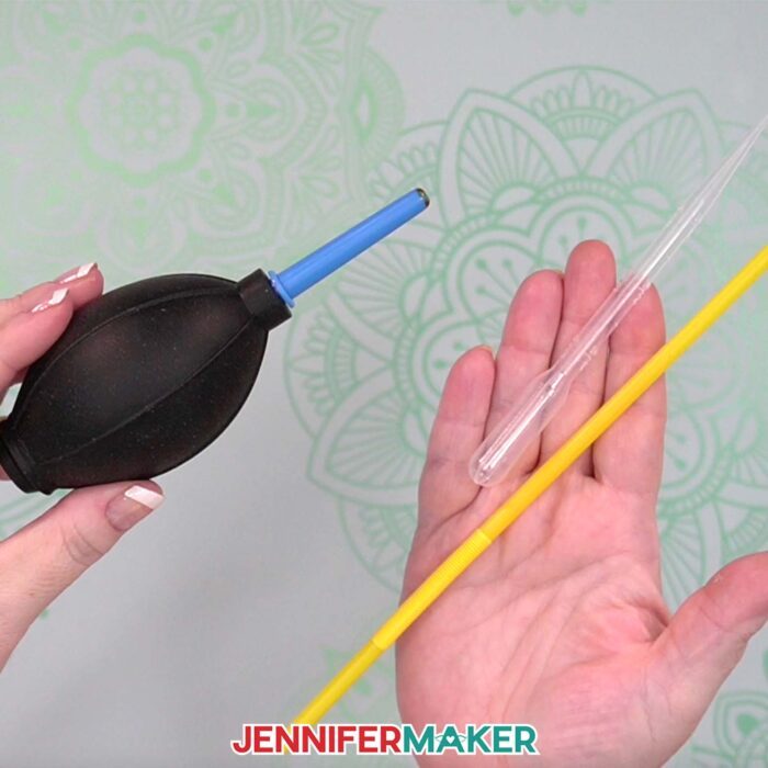 Hands show off several tools that can be used to blow Alcohol Ink On Paper for unique effects: A blower tool, a pipette, and a drinking straw